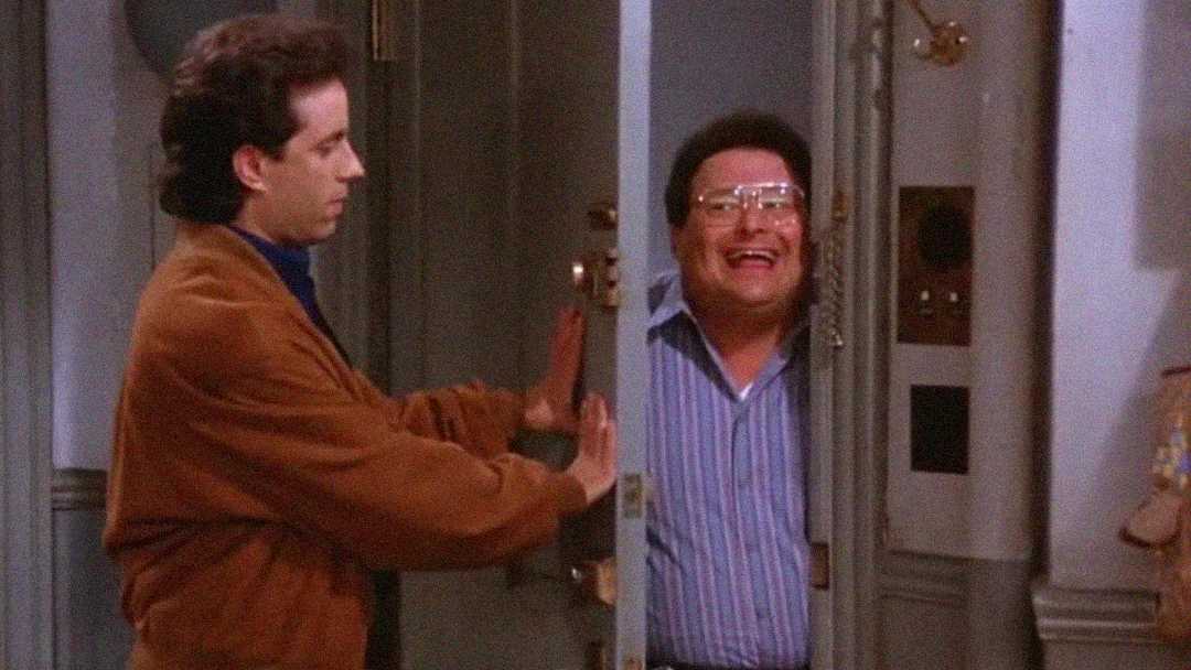 A man in a brown jacket tries to close his apartment door as another man in glasses and a striped shirt pushes it open and smiles widely from the hallway.