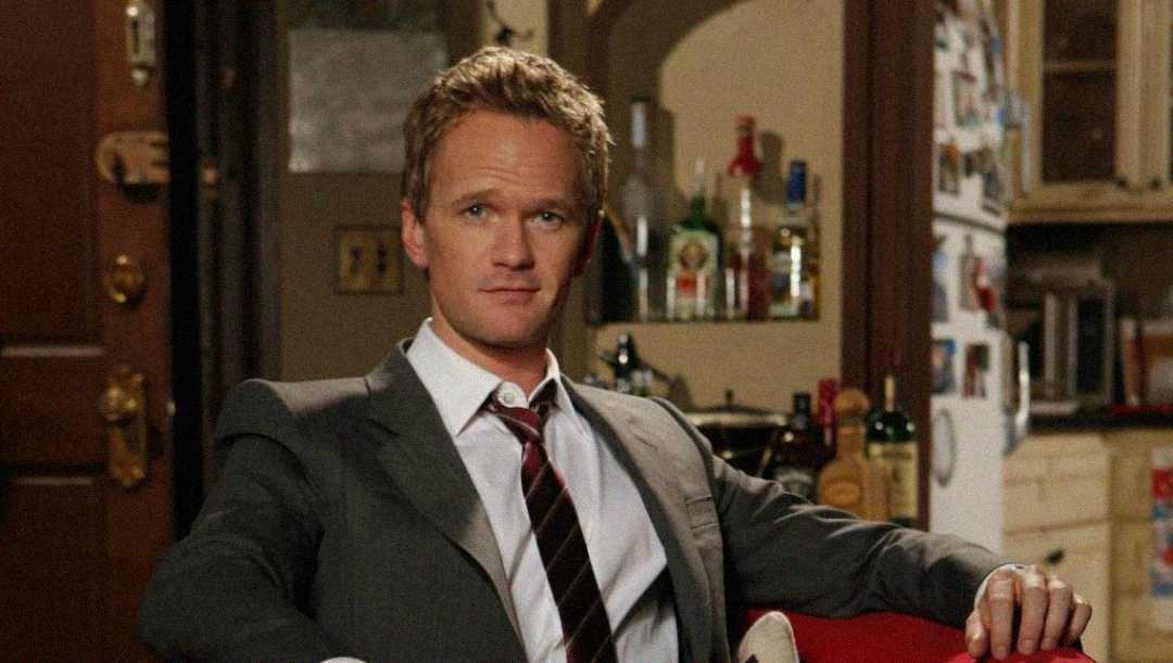 A man in a suit and tie sits on a couch in a cozy, cluttered apartment with a kitchen and shelves stocked with bottles in the background.