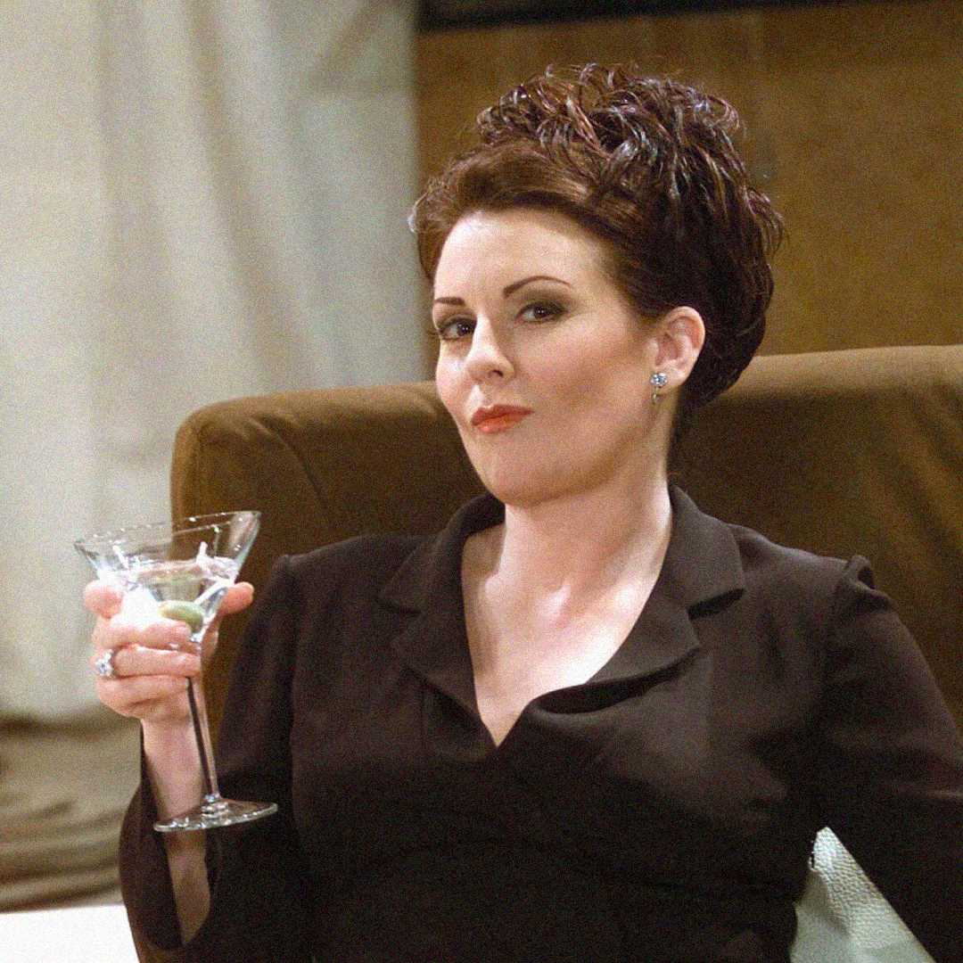 A woman with styled dark hair, wearing a dark outfit, sits on a chair holding a martini glass and looks forward with a confident, slightly amused expression.