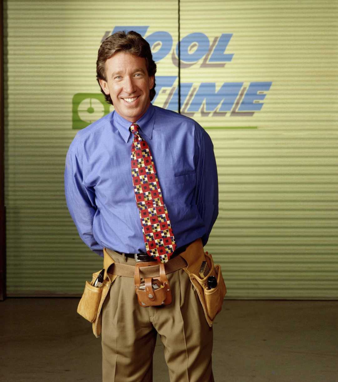 A man smiling, wearing a blue shirt, patterned tie, and tool belt, stands in front of a green garage door with the words "Tool Time" in blue letters behind him.