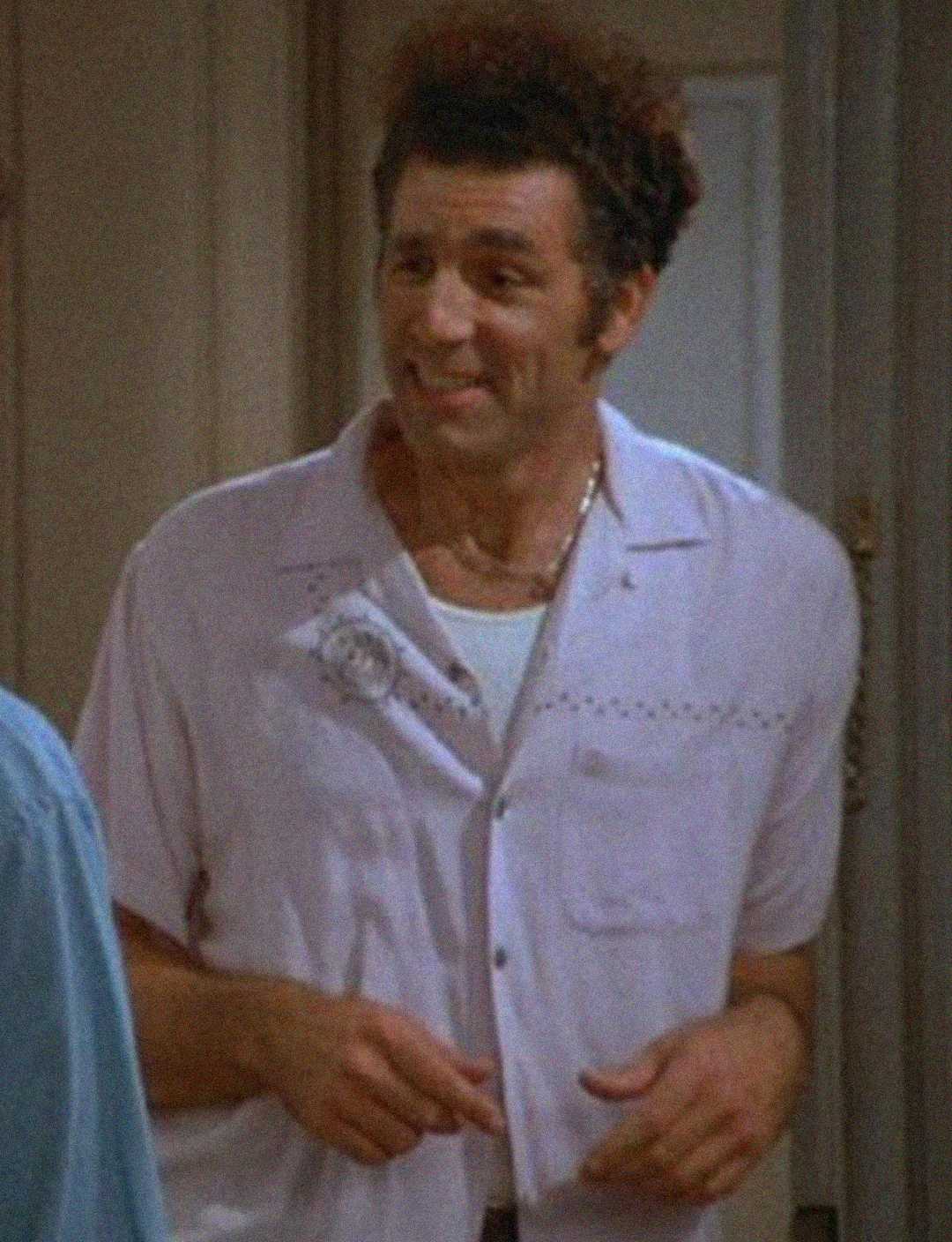 A man with dark, curly hair wearing a pale short-sleeve button-up shirt and a white undershirt smiles while standing indoors.