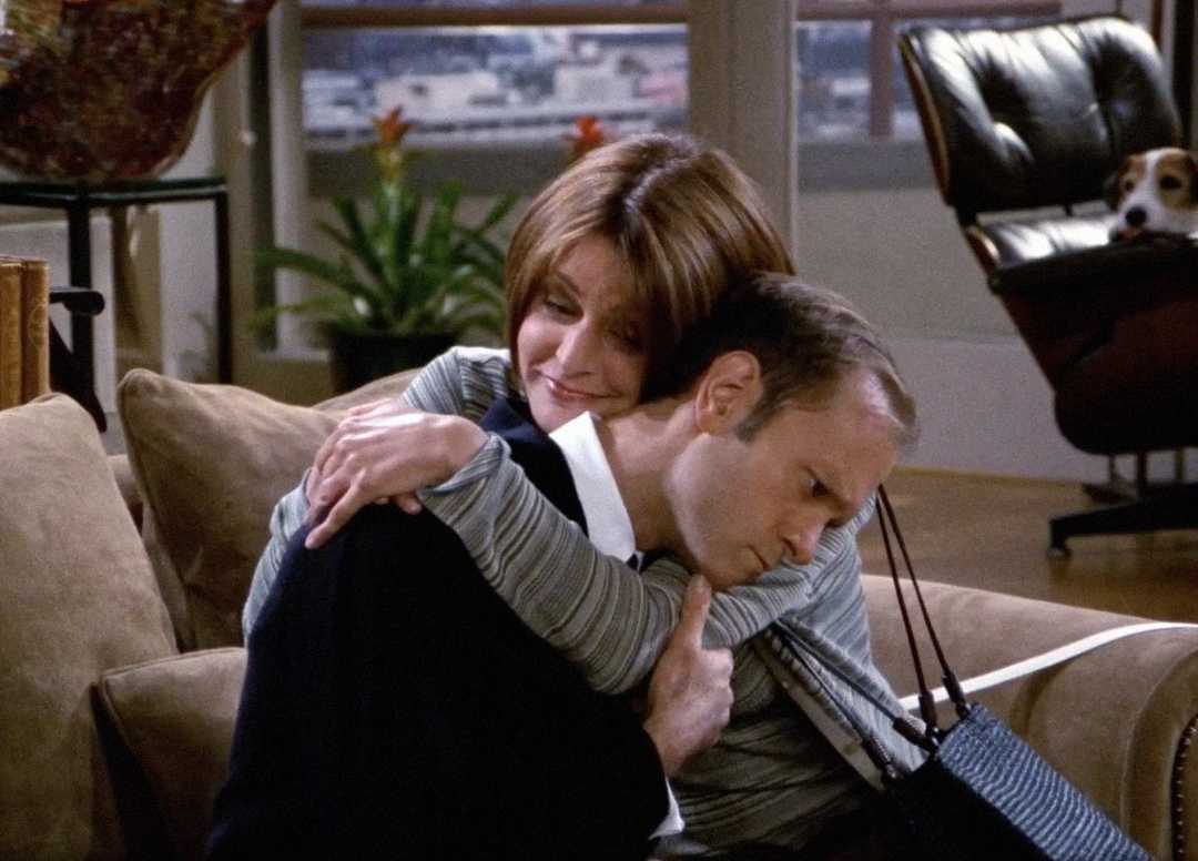 A woman with short brown hair warmly hugs a man who looks sad as they sit on a couch in a cozy living room. A dog peeks from behind a chair in the background.
