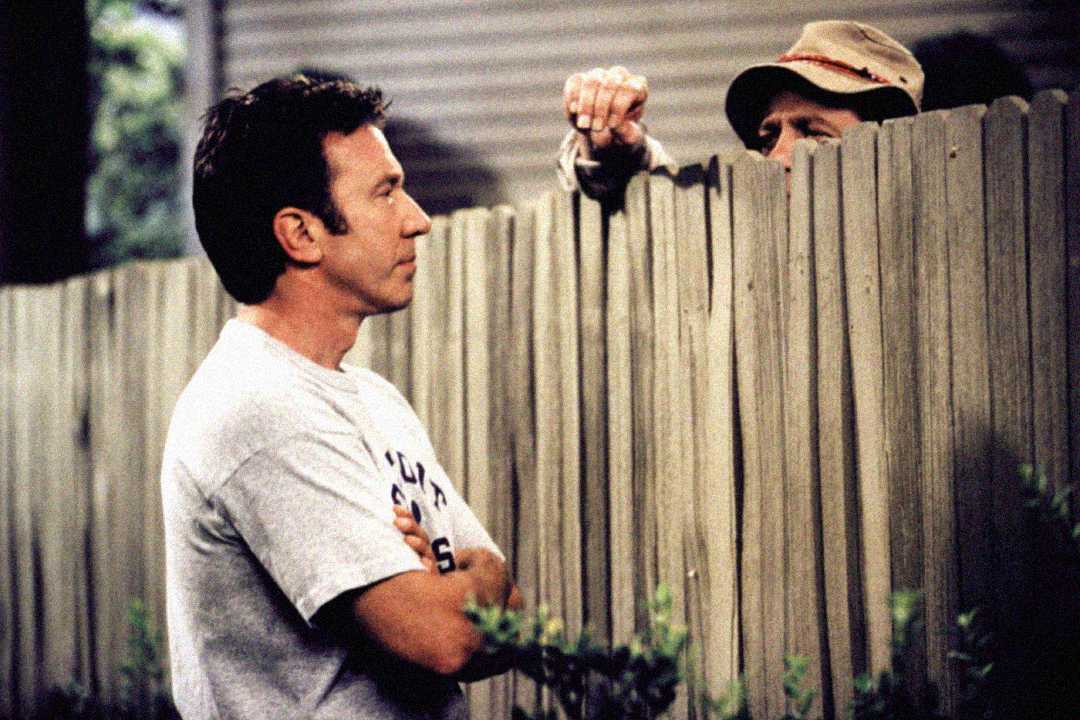 A man in a light t-shirt stands with arms crossed, talking to another man whose face is mostly hidden behind a tall wooden fence. Only the top of the second man's head and hand are visible.