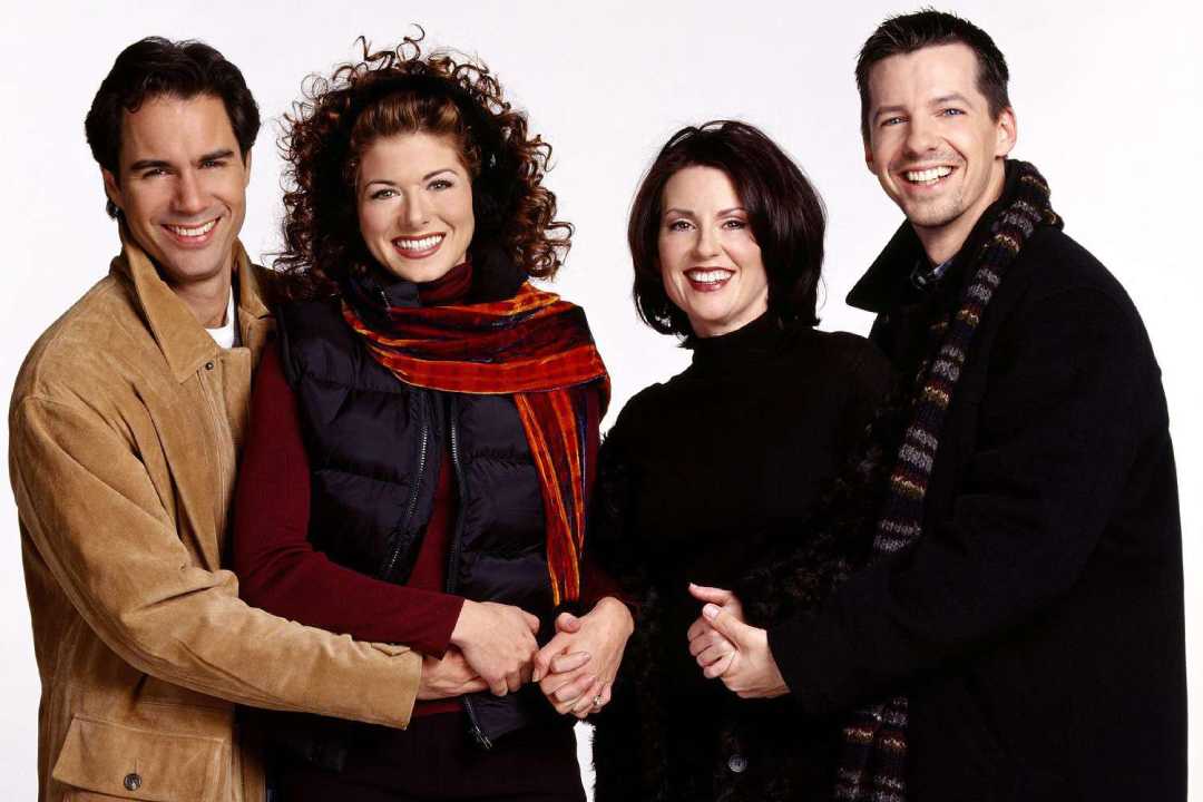 Four people stand close together smiling at the camera. Two men and two women are wearing winter clothing, including scarves, jackets, and sweaters, against a plain white background.