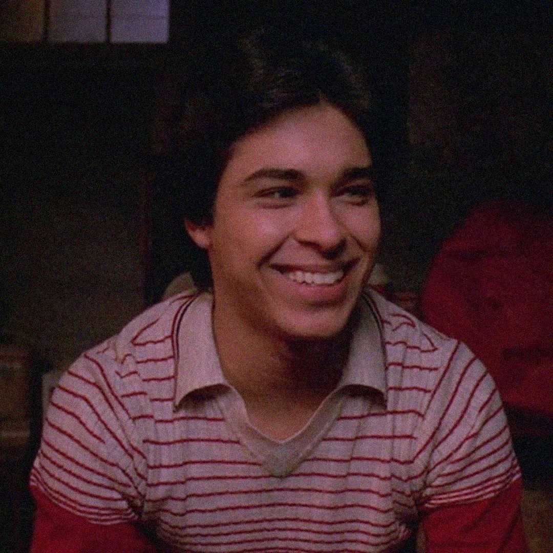 A young man with dark hair smiles warmly while sitting indoors. He is wearing a white and red striped sweater over a collared shirt. The background is dimly lit.