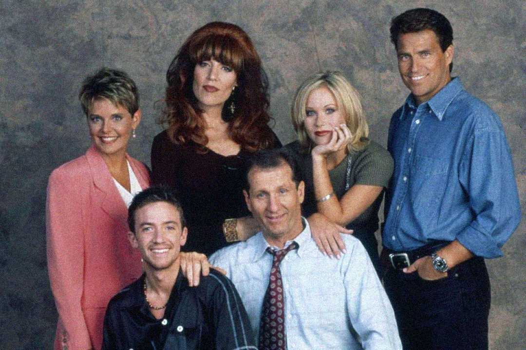 A group of five people, three women and two men, pose together in front of a neutral backdrop. They appear to be actors from a sitcom, standing and sitting closely, smiling and looking at the camera.