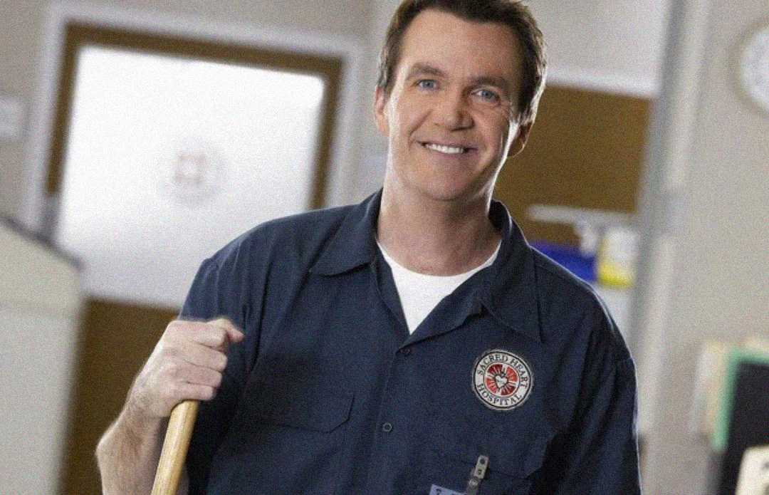 A smiling man in a blue janitor uniform holds a mop in what appears to be a hospital or clinic hallway.