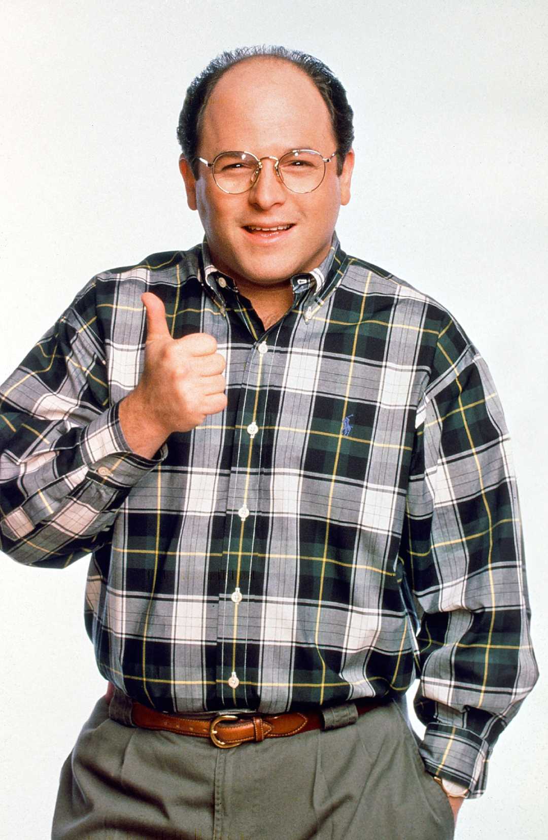A man with glasses and a receding hairline wears a black, white, and green plaid shirt and khaki pants. He is smiling and giving a thumbs-up gesture against a plain light background.