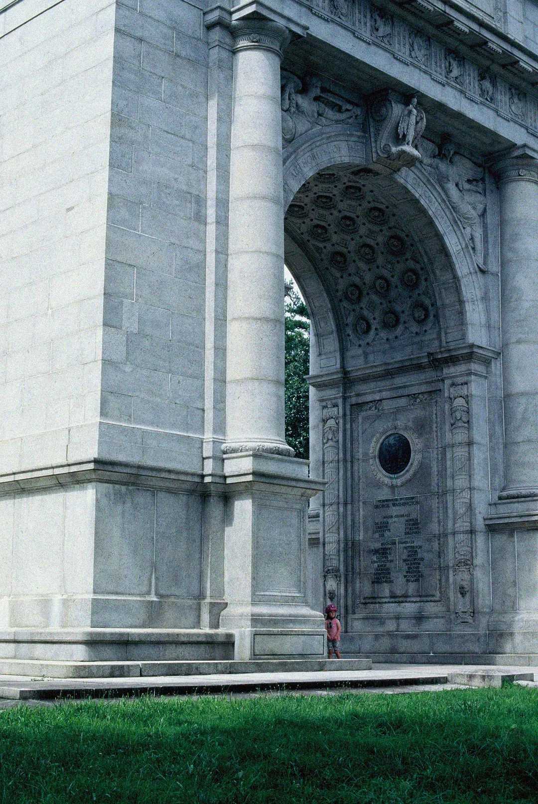 A small child in pink stands near the base of a large, ornate stone arch monument with carved details, surrounded by green grass.