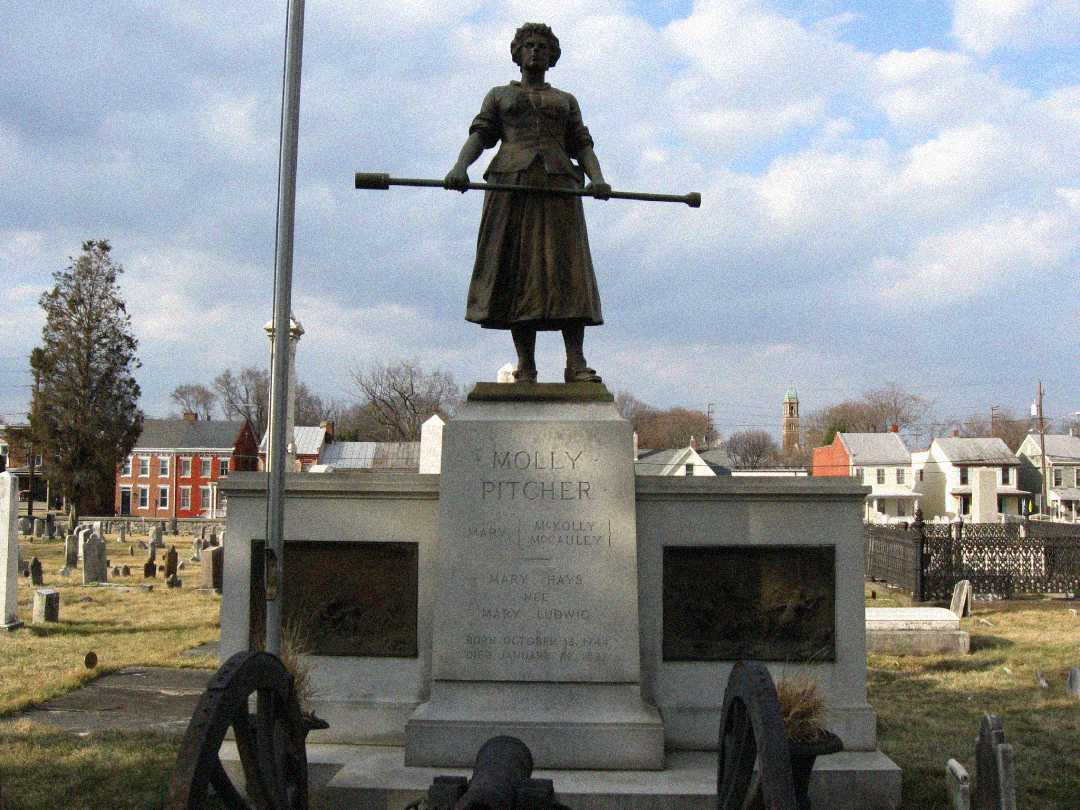A statue of a woman holding a cannon ramrod stands atop a stone monument labeled "Molly Pitcher," surrounded by cannons and gravestones in a cemetery with houses in the background.
