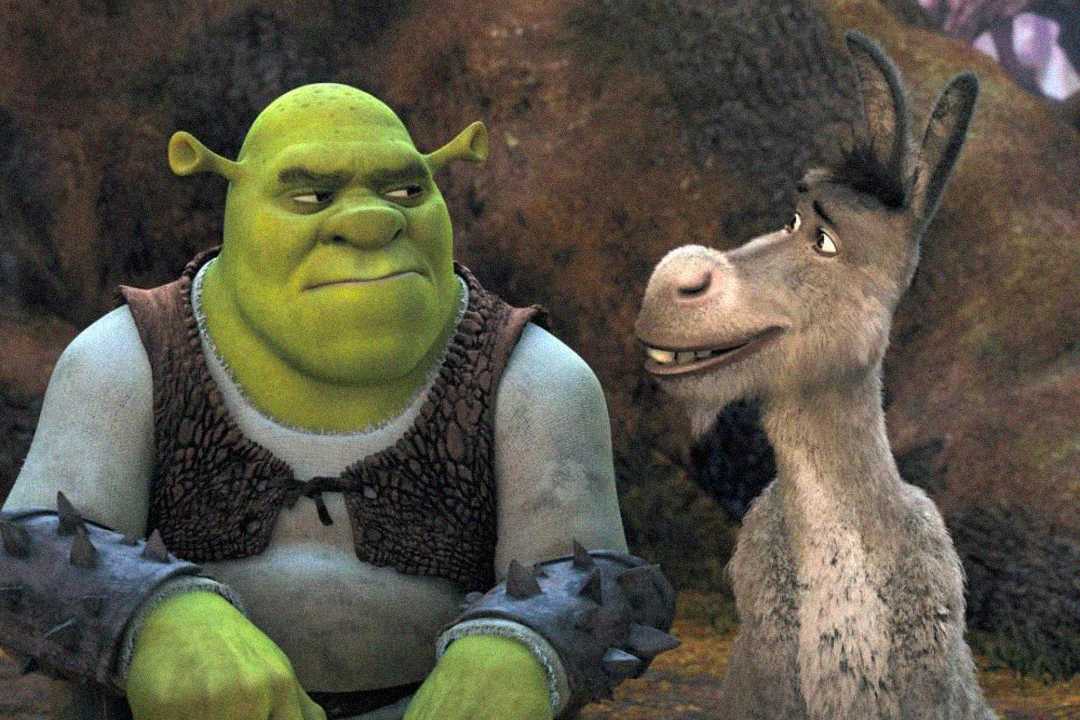 Shrek, a large green ogre, sits next to Donkey, his grey donkey friend, in a forest setting. Shrek looks slightly annoyed while Donkey smiles and talks to him.