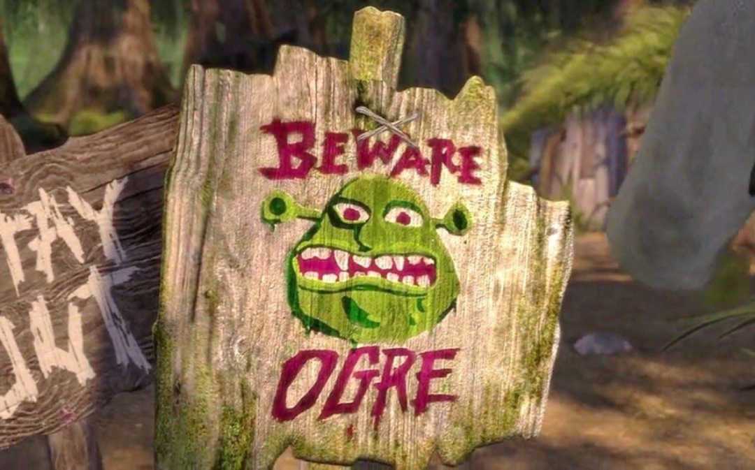 A wooden sign with a painted green ogre face and bold red text that reads "BEWARE OGRE." The sign is nailed to a tree and appears weathered, set in a forest background.