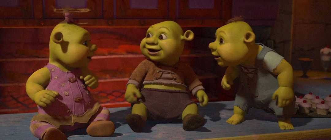 Three animated ogre babies with green skin sit together on a blue floor, facing each other, wearing different colored outfits. The background shows a warm, indoor setting with cupcakes on the right side.