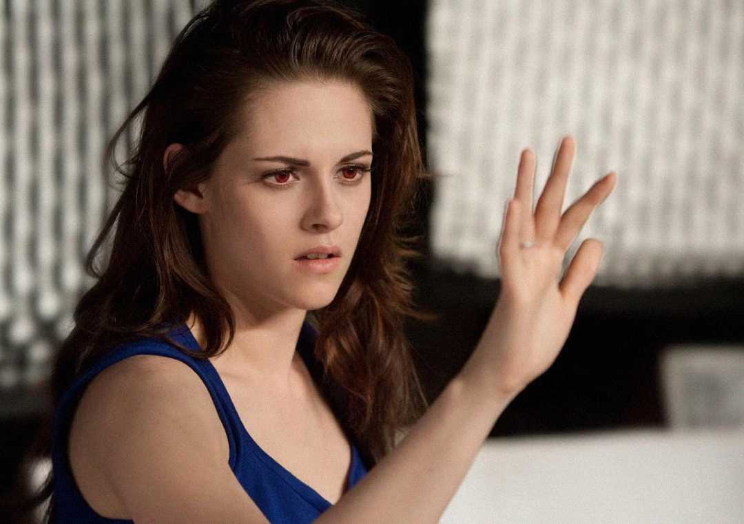 A woman with long brown hair wearing a blue top looks intently at her raised hand, with a serious expression and red eyes, against a blurred background.