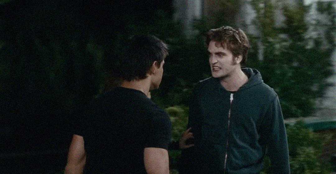 Two young men stand outside at night. One, wearing a dark hoodie, looks angry and is yelling at the other, who has short dark hair and wears a black T-shirt. The background is dimly lit with greenery.