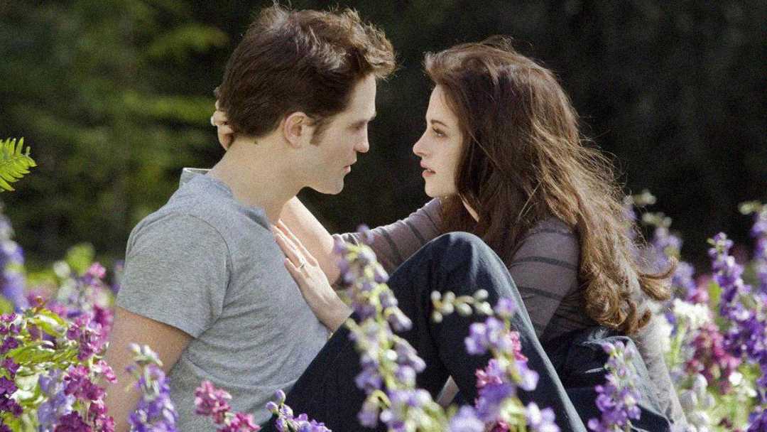A young couple sits closely together in a field of purple flowers, gazing into each other's eyes. The woman has long brown hair and touches the man's neck lovingly; he wears a gray t-shirt. The mood is intimate and gentle.