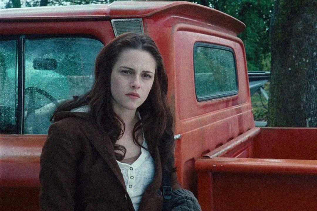 A young woman with long brown hair stands next to an old red pickup truck on a rainy day, looking serious and thoughtful. She wears a brown jacket over a white shirt. Trees and other cars are in the background.