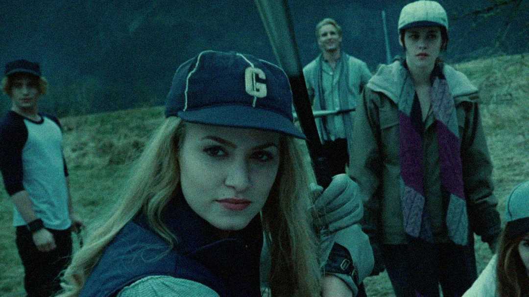 A young woman in a baseball cap with a “G” on it holds a bat, looking at the camera, surrounded by four people in outdoor gear on a grassy field with mountains in the background. The scene is dimly lit and moody.