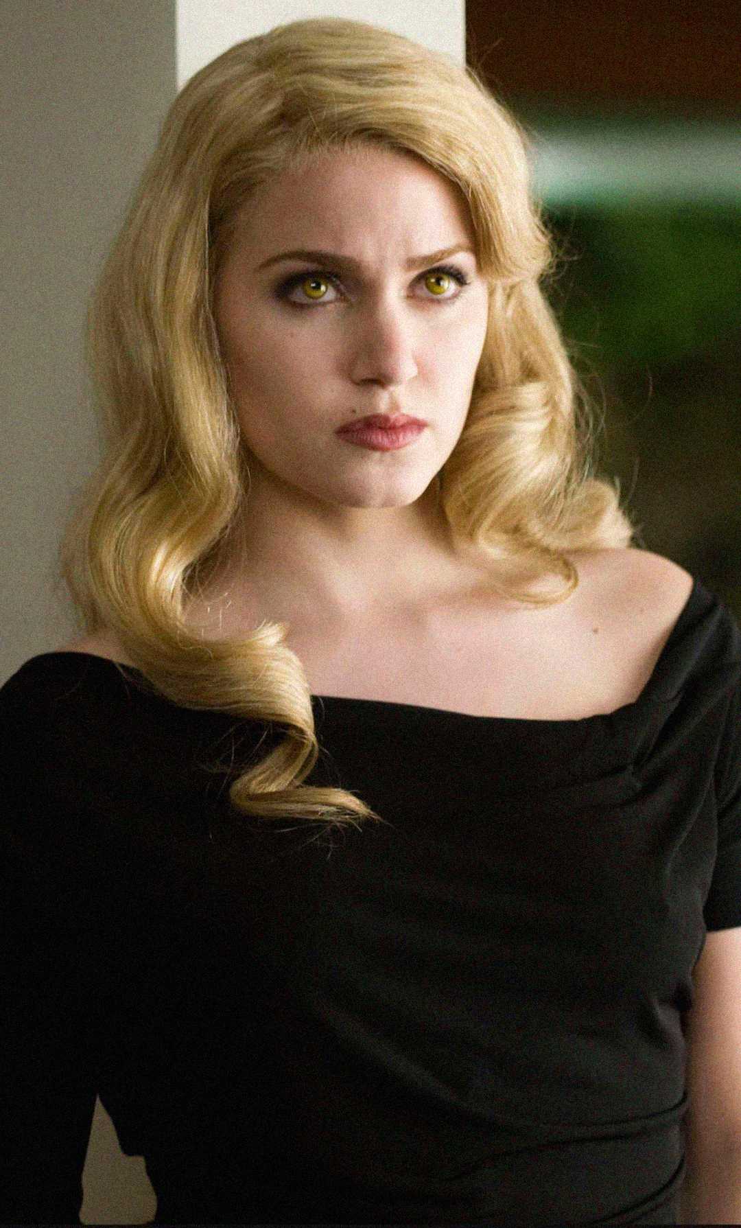 A woman with long, wavy blonde hair and striking golden eyes wears an off-the-shoulder black top, standing indoors and looking serious.