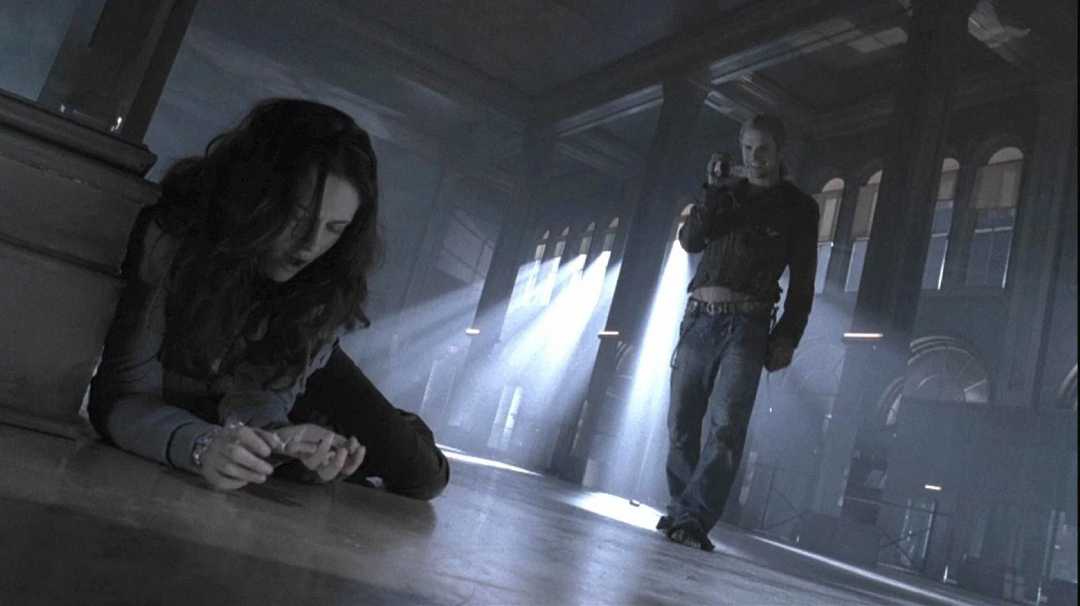 A woman is lying on the floor in a large, dimly lit hall, looking down. Behind her, a person holding a gun is walking toward her through beams of light streaming from tall windows.