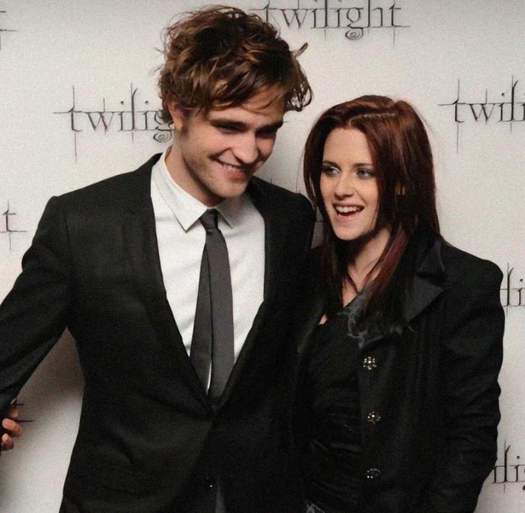 A man in a suit and tie and a woman in a black outfit smile together in front of a white backdrop with “Twilight” written on it multiple times.