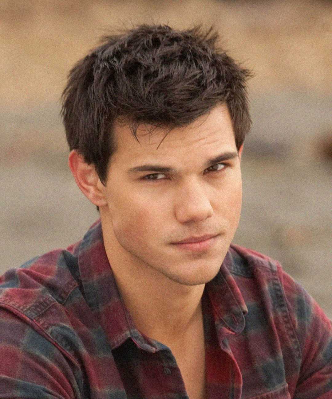 A young man with short dark hair and a slight smile, wearing a red and black plaid shirt, looks directly at the camera against a blurred outdoor background.