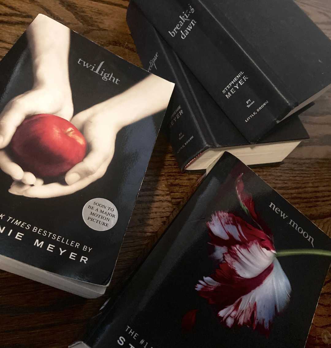Four Twilight Saga books by Stephenie Meyer are arranged on a wooden surface. The covers of “Twilight,” “New Moon,” “Eclipse,” and “Breaking Dawn” are visible, featuring their distinct cover art.