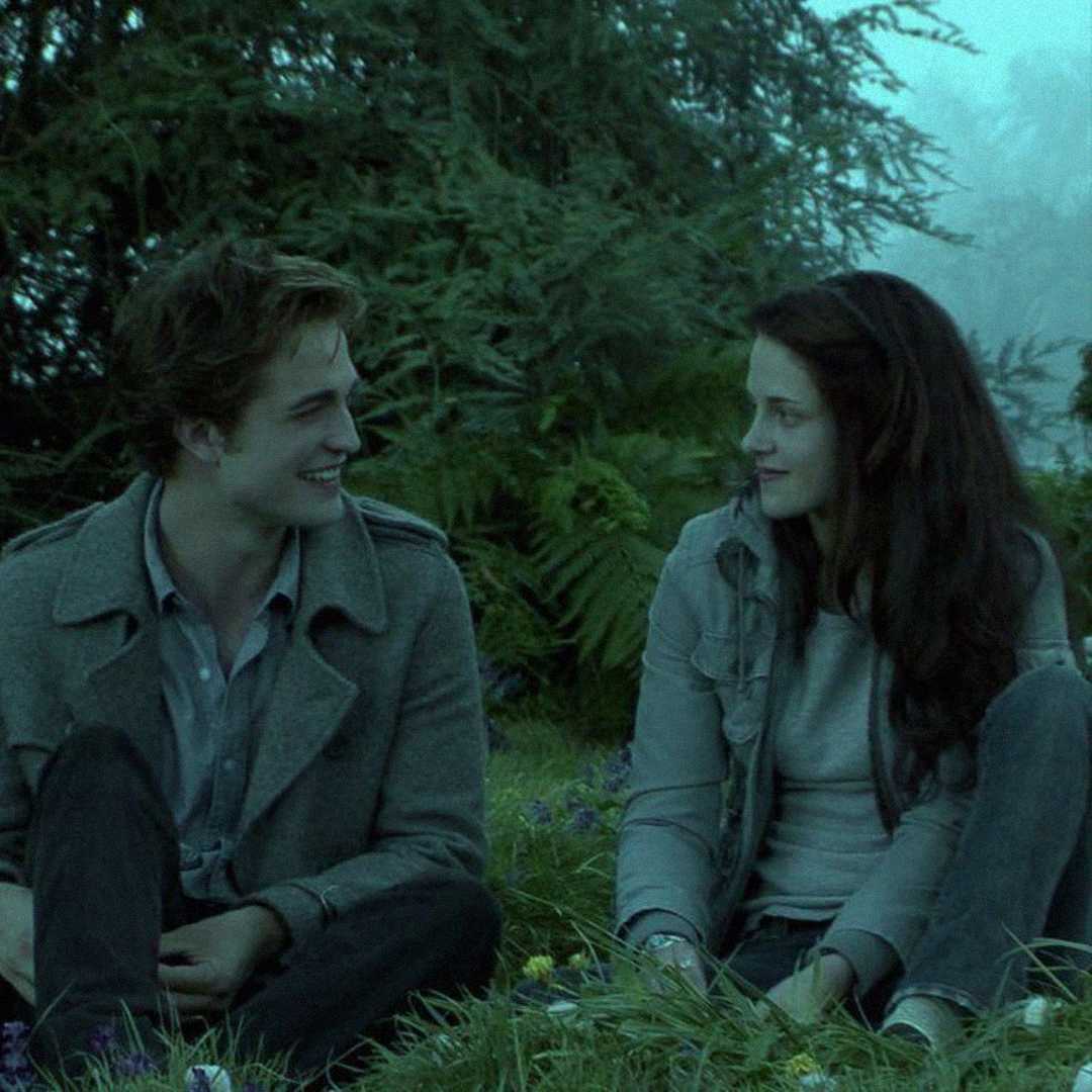 A young man and woman sit on grass in a forest clearing, facing each other and smiling, with green trees in the background and soft, muted lighting.