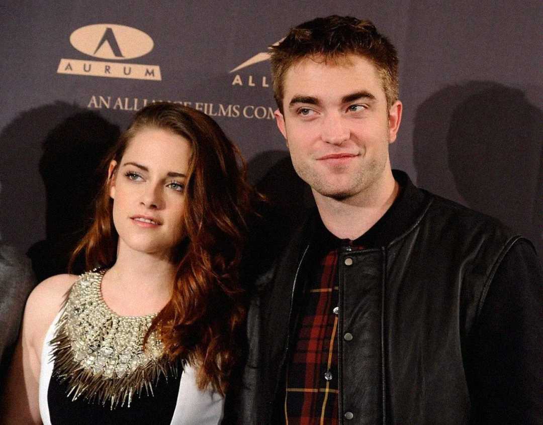 Two people pose together at an event, standing in front of a dark backdrop with logos and gold text. The woman wears a decorative sleeveless dress, and the man wears a plaid shirt and black jacket. Both look slightly off camera.