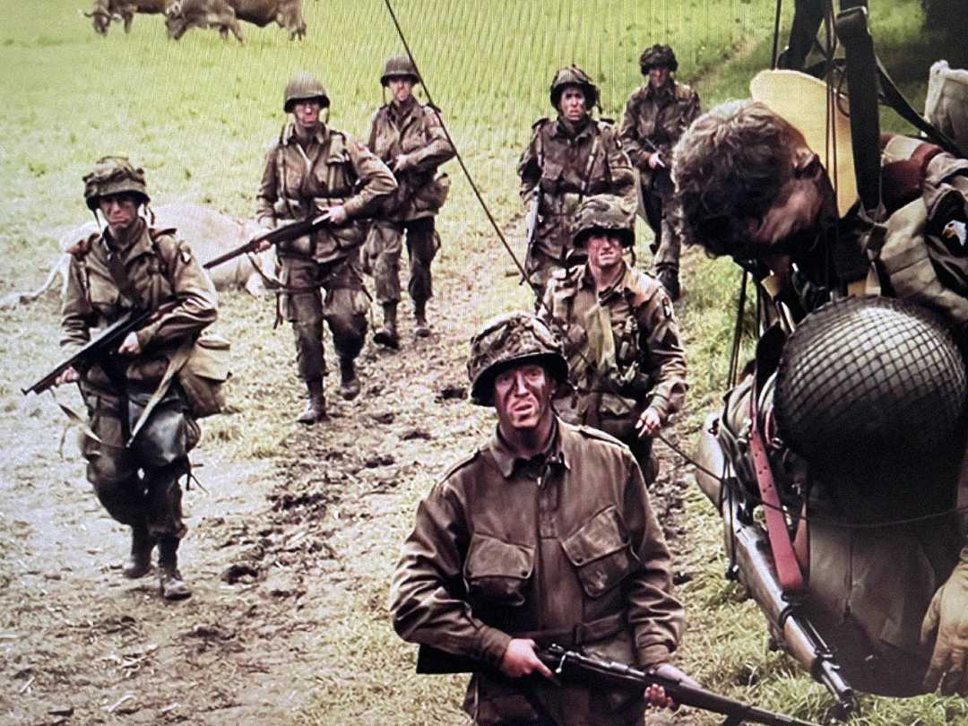 A group of soldiers in World War II-era uniforms walk through a muddy field, armed with rifles and wearing helmets, with one soldier in the foreground looking fatigued.