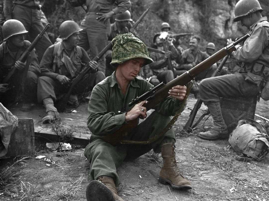 A soldier in green uniform and camo helmet sits on the ground inspecting his rifle, while other soldiers with helmets and rifles sit and stand in the background, appearing to rest in a wooded outdoor area.