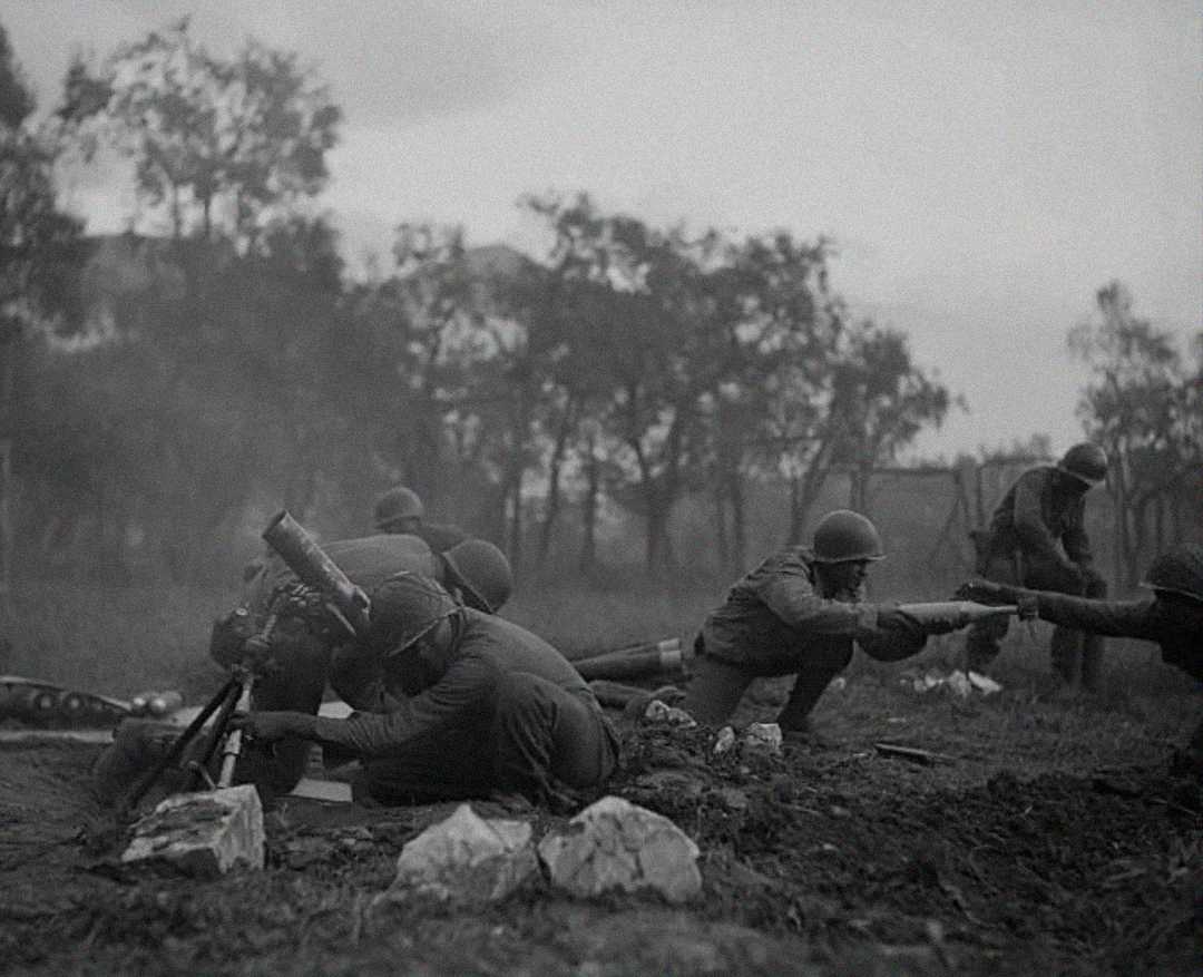 Soldiers in helmets operate a mortar and assist each other in a muddy field, surrounded by rocks and trees, during a battle in a smoky, overcast environment.