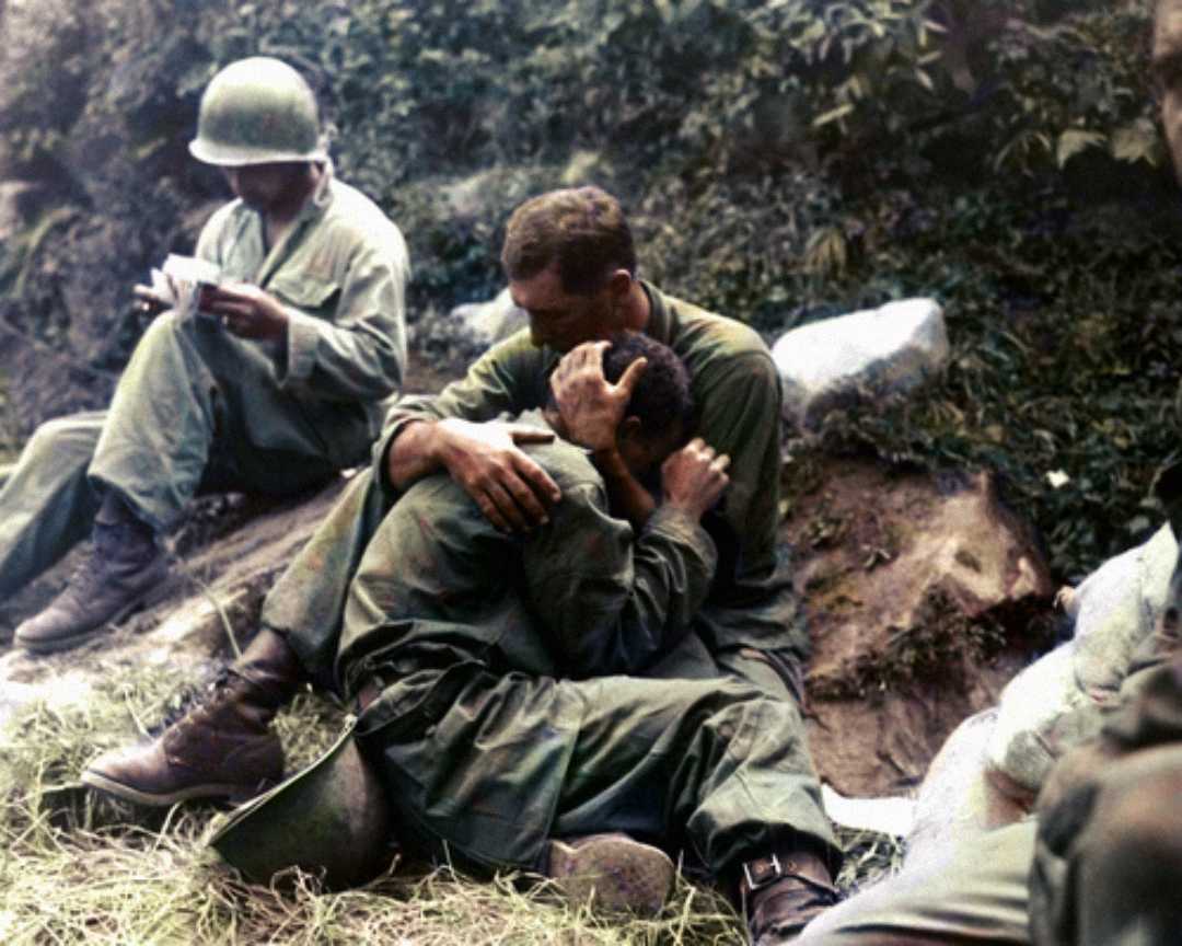 A soldier sits on the ground, holding and comforting a distressed comrade who is leaning into his chest. Another soldier nearby reads a letter, with greenery and rocks in the background.