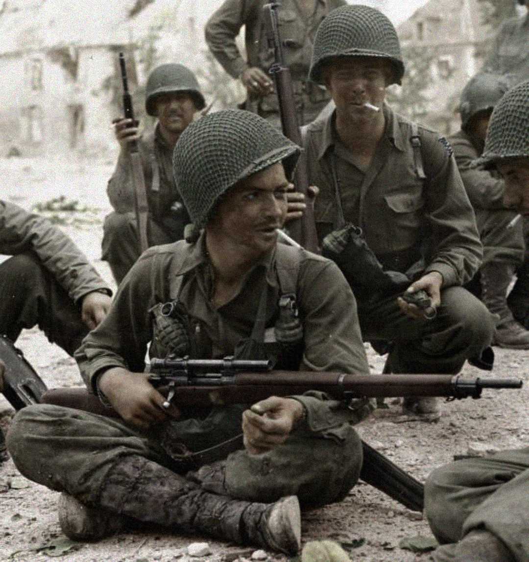 A group of World War II soldiers in uniform and helmets sit and crouch on the ground, some smiling and holding rifles, with buildings visible in the blurred background.