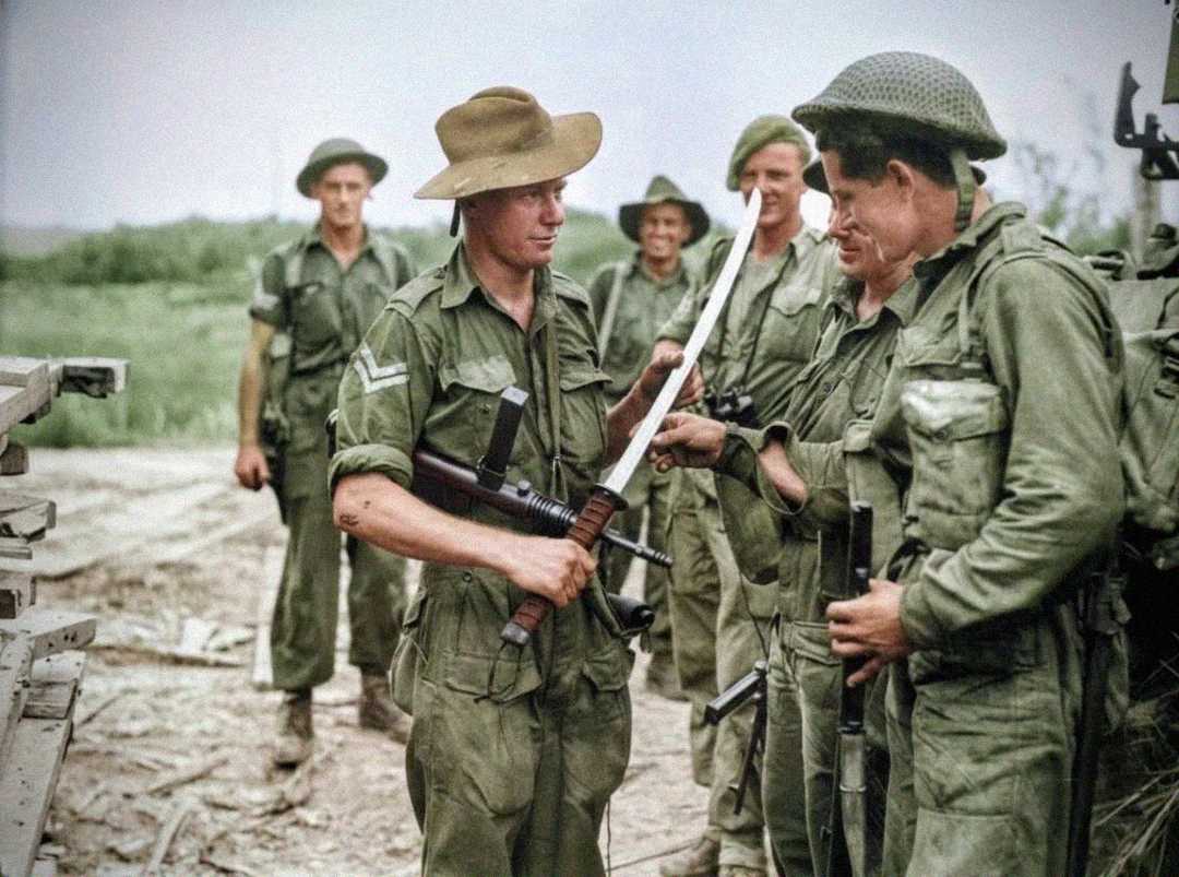 A group of World War II soldiers in green uniforms stand outdoors, smiling and examining a long machete. One soldier holds a machine gun and the machete, while others wear helmets and hats, watching with interest.