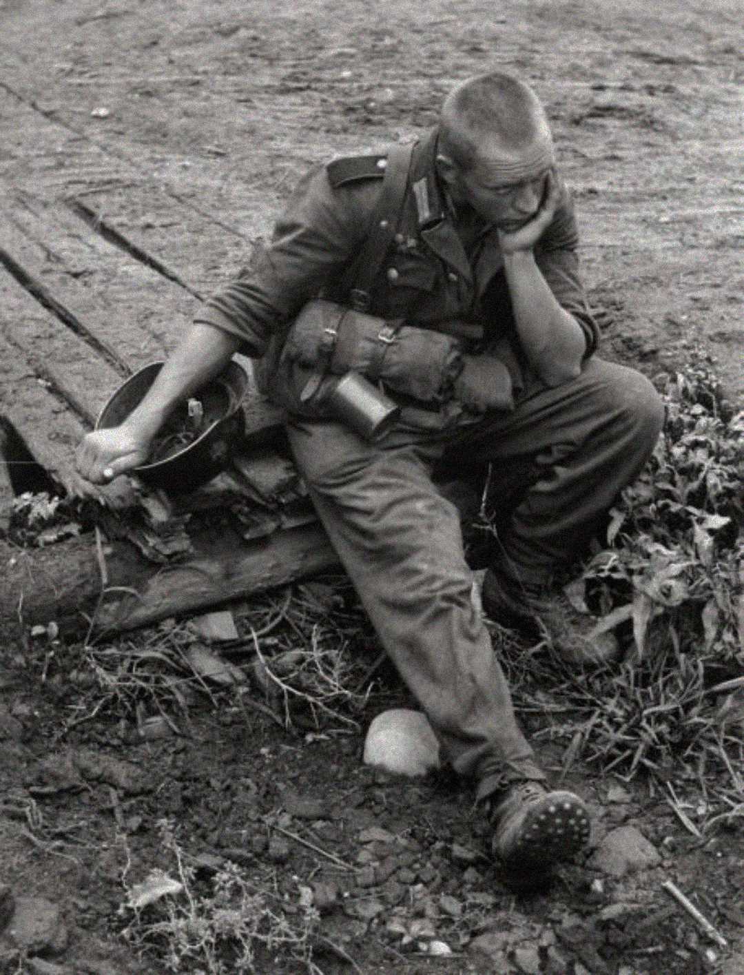 A soldier in uniform sits on a log, head resting on his hand in a thoughtful pose. He appears tired, with his helmet and gear beside him on the ground. The setting is outdoors, with bare earth and scattered plants.