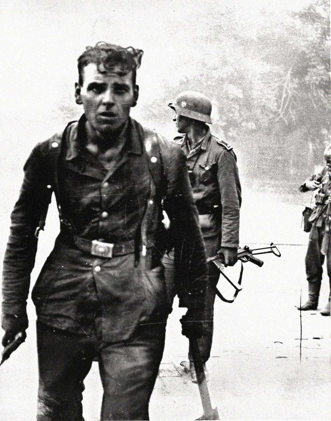 A weary soldier walks toward the camera holding a weapon, while two other soldiers in military uniforms and helmets, one holding a submachine gun, stand behind him in a smoky, war-torn environment.