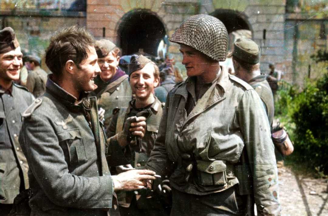 A group of soldiers, some smiling, interact outdoors. One soldier in a netted helmet shakes hands with another in a uniform. Stone archways and greenery are visible in the background.