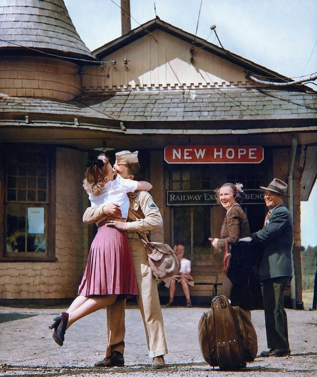 A soldier in uniform passionately kisses a woman who has leapt into his arms outside a train station. Two older people smile nearby, with luggage in the foreground and a sign reading "New Hope" above them.