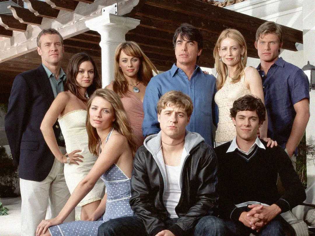 Ten people, dressed in stylish casual and semi-formal clothing, pose together outdoors in front of a house with white pillars and wooden beams. Some are seated, others standing, all looking toward the camera.