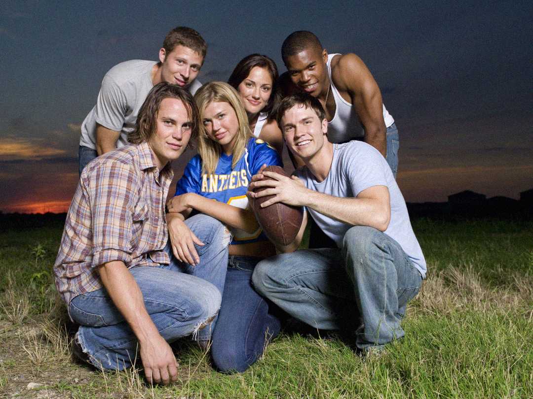 Six young adults pose together in a grassy field at sunset; one woman wears a blue football jersey, and one man holds a football. They all smile and lean closely for the photo.