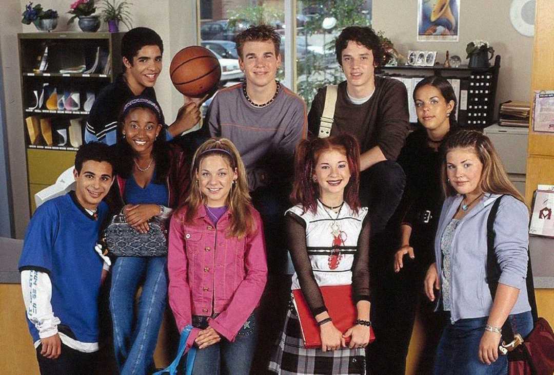 A group of nine teenagers pose together in a school setting, smiling at the camera. Some are standing, others are sitting on a desk. One holds a basketball; books and folders are visible in the background.