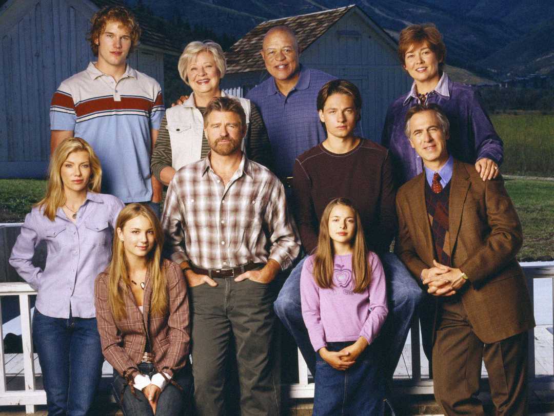 A group of eleven people, including men, women, and children, are posing outdoors in front of a white fence and barn, with mountains in the background. They are all smiling and dressed in casual clothing.