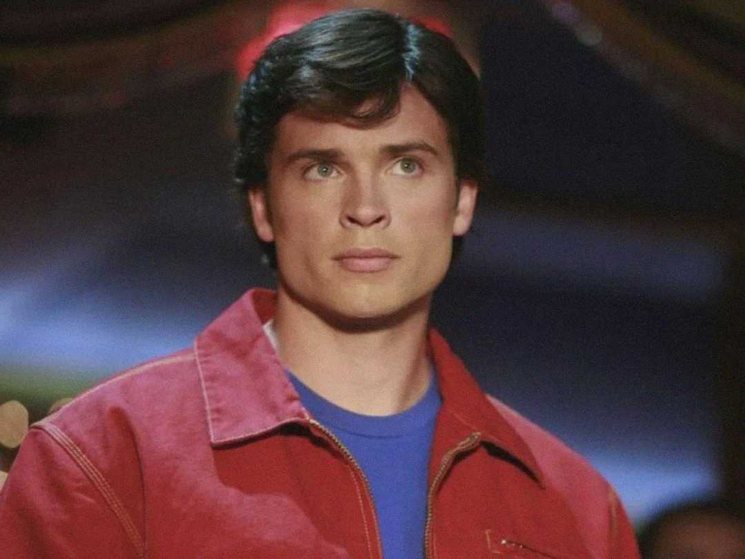 A young man with short brown hair and green eyes wears a red jacket over a blue shirt, looking serious in a softly lit indoor setting.