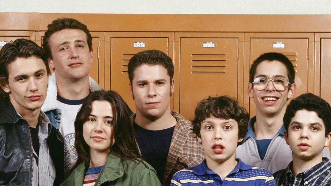 Seven teenagers stand in front of orange school lockers, posing for a group photo. They wear casual clothes and various expressions, ranging from serious to smiling.