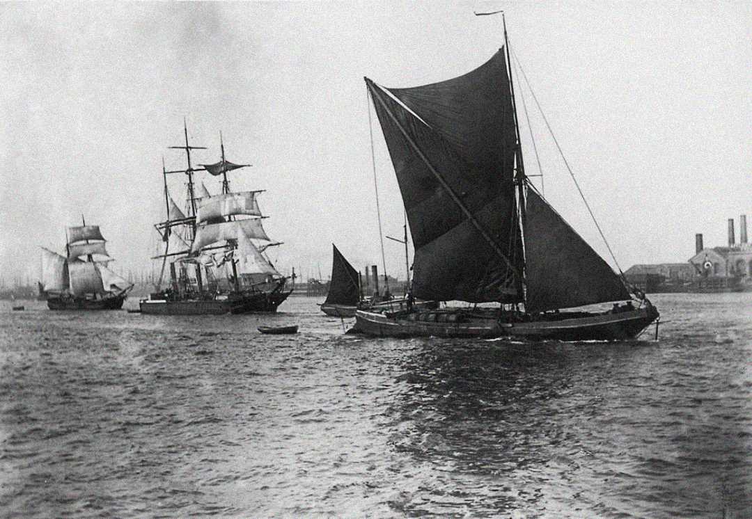 Historic black-and-white photo of several large sailing ships and smaller boats on a river, with industrial buildings and smokestacks visible in the background under a cloudy sky.