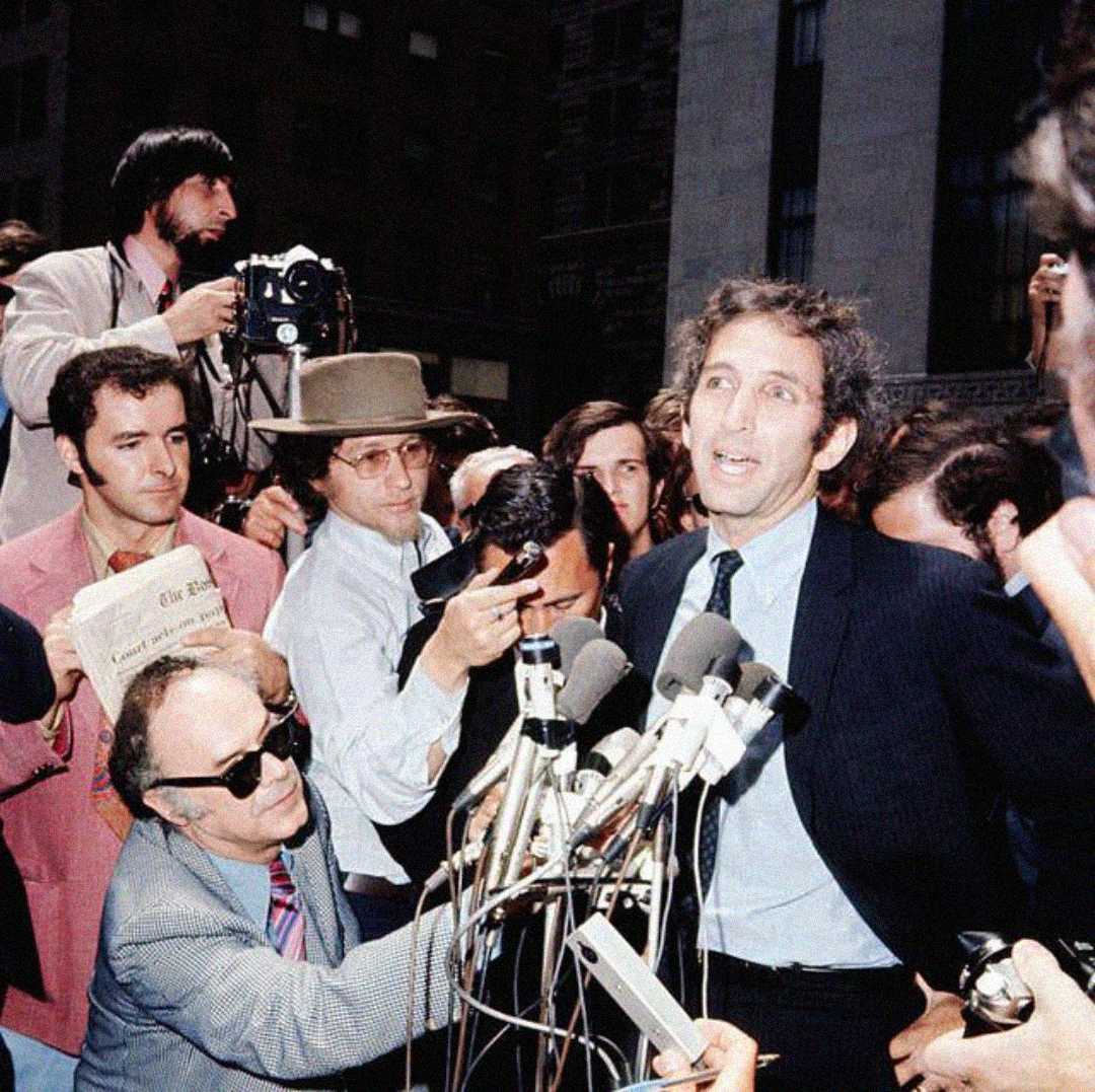 A man in a suit speaks into multiple microphones outside, surrounded by reporters, photographers, and cameramen, as a crowd gathers behind them.