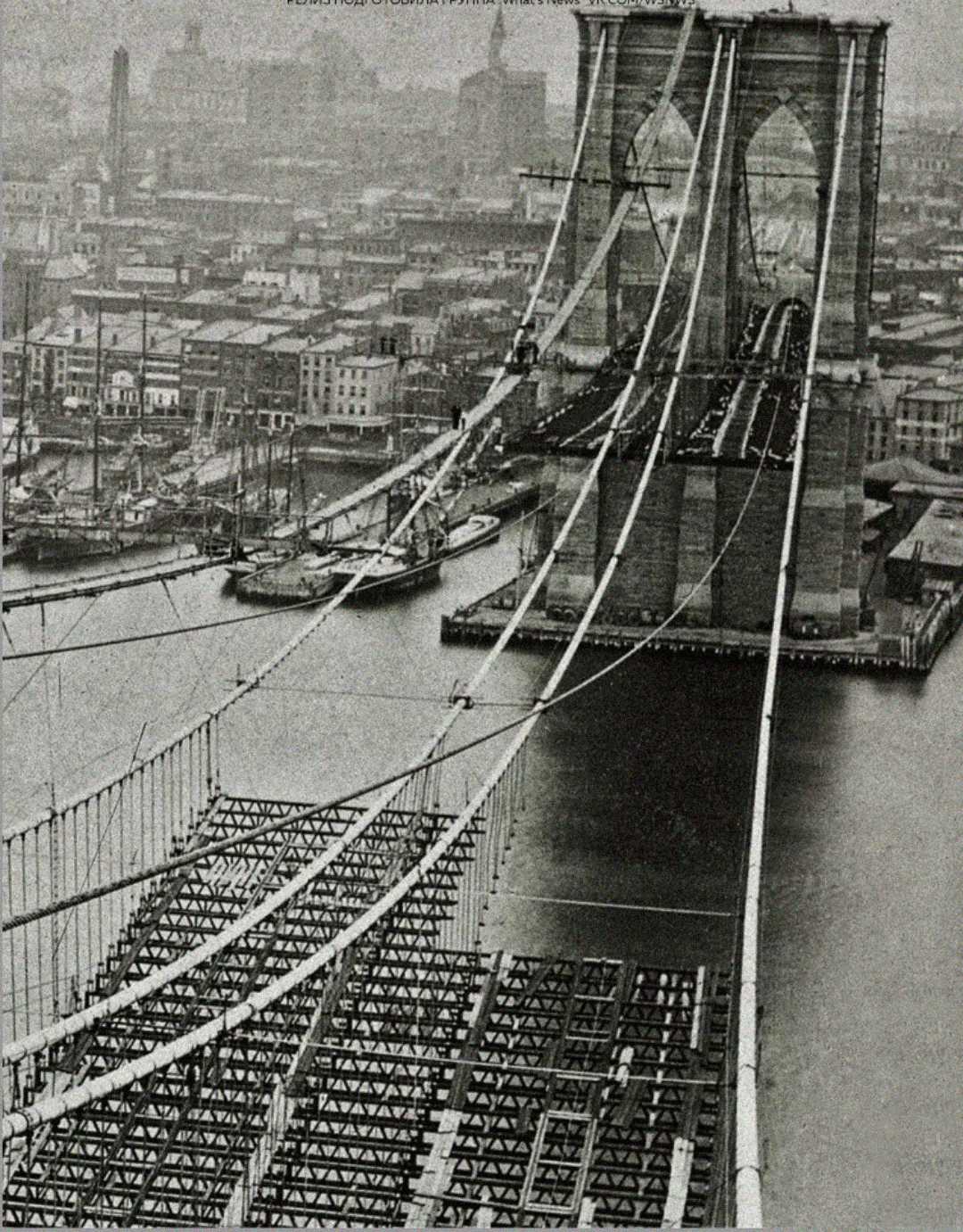 A black-and-white photo shows the Brooklyn Bridge under construction, with cables and incomplete roadway spanning the East River and the New York City skyline visible in the background.