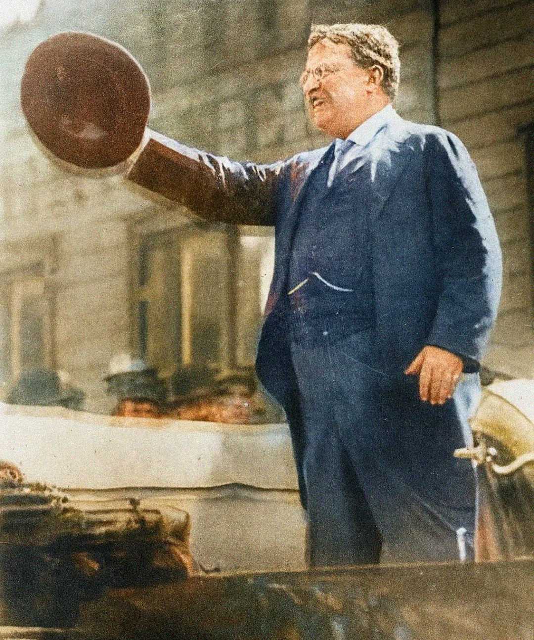 A man in a suit smiles and waves a round hat in the air while standing outdoors, likely addressing a crowd. The background shows blurred people and building facades. The image has a historic, vintage look.