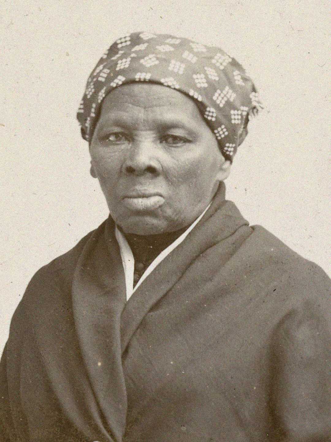 An older Black woman wearing a patterned headscarf and a dark shawl looks directly at the camera with a serious expression, in a sepia-toned historical portrait.